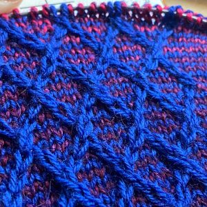 Close up of a sock in progress in pink, blue, and purple. A cable pattern crosses over and over again on the front of the sock.
